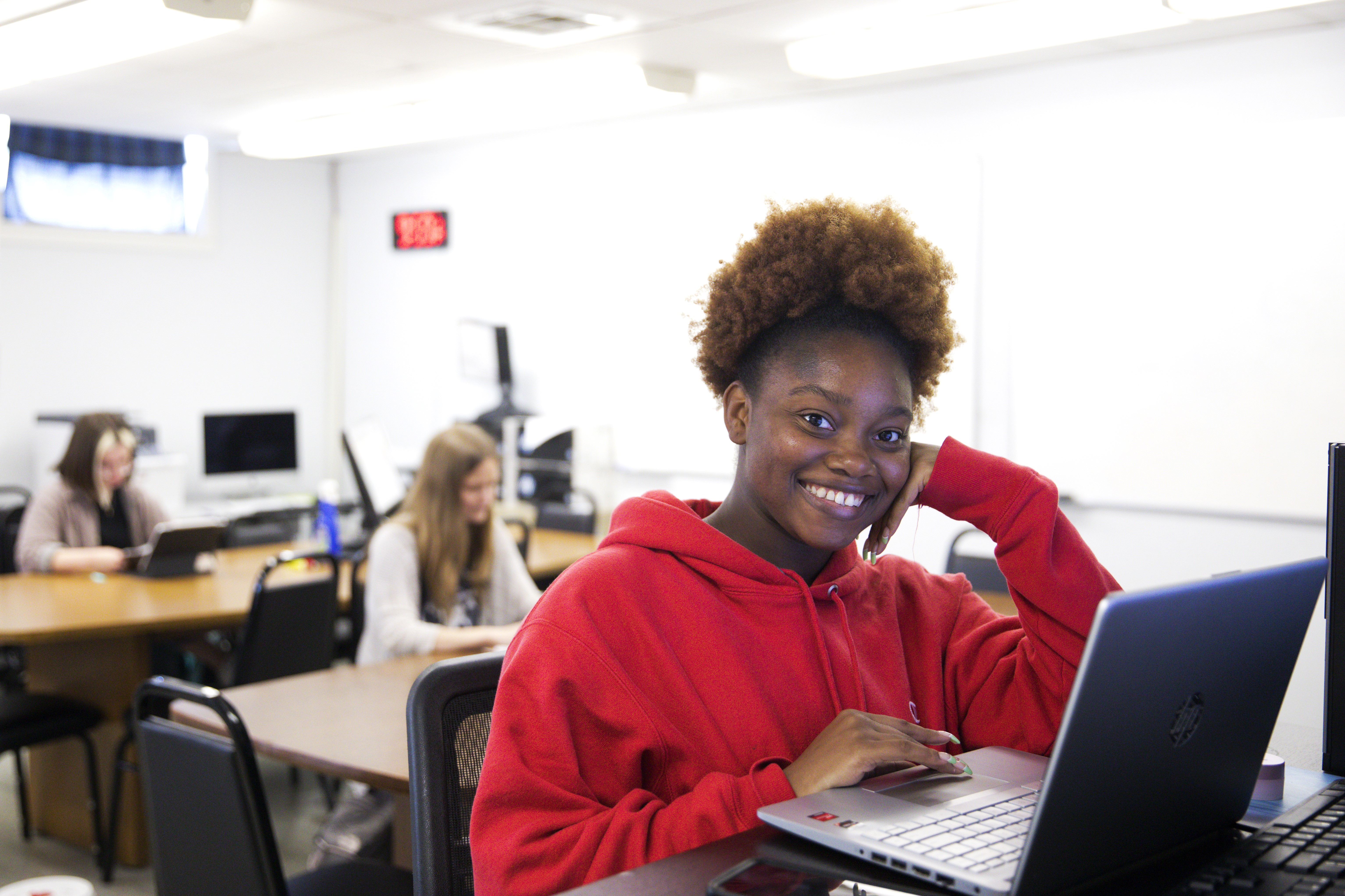 Student wearing red sweatshirt smiles at camera in front of laptop. Two other workers are seated in the background